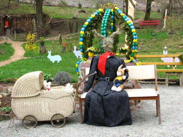 Die Osterhasenfrau mit dem Osterhasennachwuchs im Kinderwagen ruht auf der Bank vor dem geschmückten Osterbrunnen